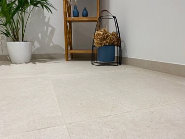 Close-up van vloer met Sevilla Cream keramische tegels in kalksteenlook. Een houten plank, een plant en decoraties op de achtergrond