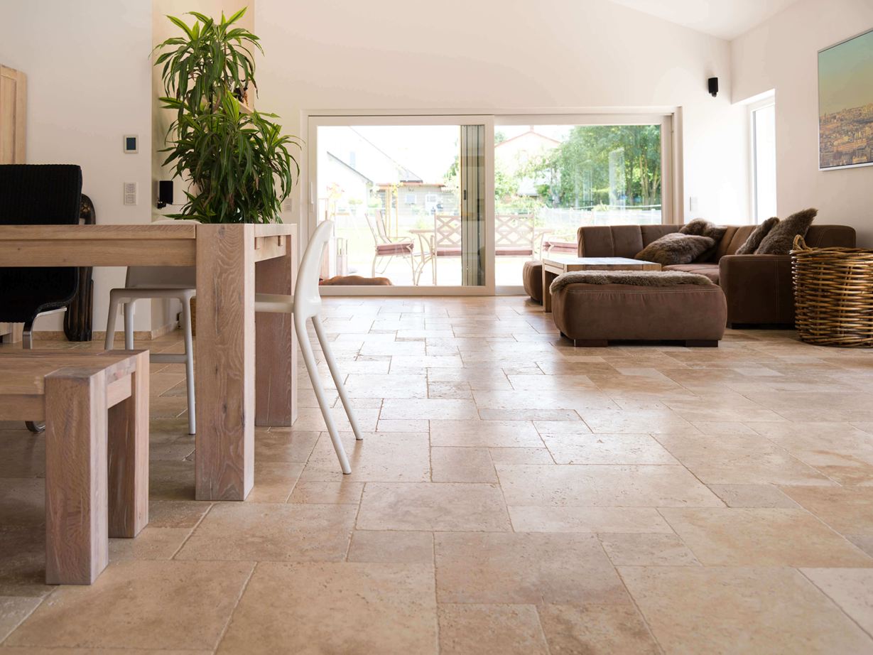 Die hellen Farbtöne wirken leicht und elegant. Woonkamer met bruine bank, witte muren en medium travertin tegels in bruin-beige als vloerbedekking