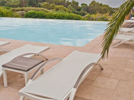 Witte ligbedden op een travertijnen vloer bij een zwembad, een palmtak steekt van rechts in het beeld