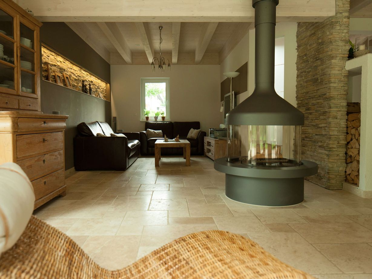 Woonkamer met vrijstaande open haard en zithoek op Lichte travertin tegels, een rieten fauteuil op de voorgrond