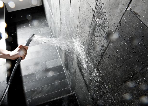 Hand houdt een douchekop tegen een muur met donkere stenen tegels